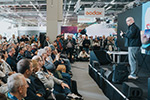 Publikum bei einer Live-Präsentation auf der Messe, aufmerksam zuhörend vor einer Bühne mit Referent.